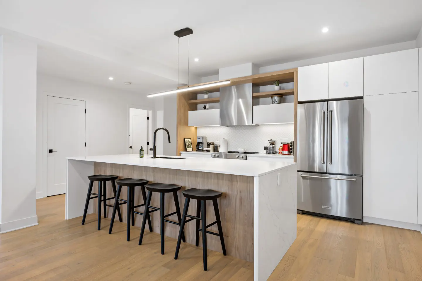 Bright kitchen with white cabinets, stainless steel fridge, and island with black stools. Wood floors and modern lighting.