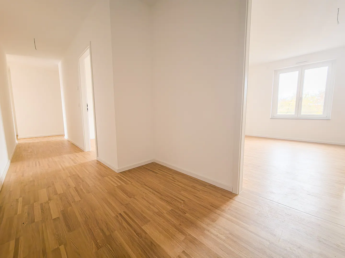 Bright, empty room with light wood floors and white walls. A window is visible in the adjacent room.
