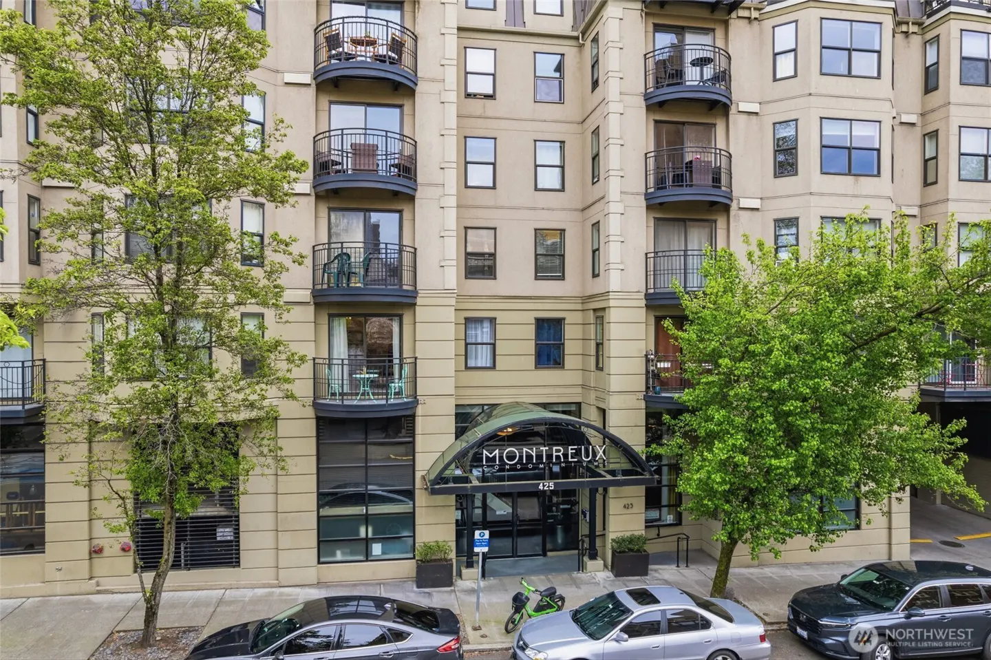 Exterior view of the Montreux Condominiums, a beige building with black-railed balconies and a glass-covered entrance. Cars are parked on the street.