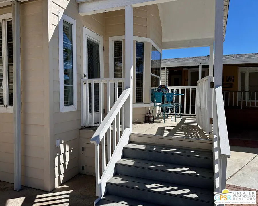 Exterior view of a beige house with white trim, featuring a porch with blue patio furniture. Steps lead up to the porch.