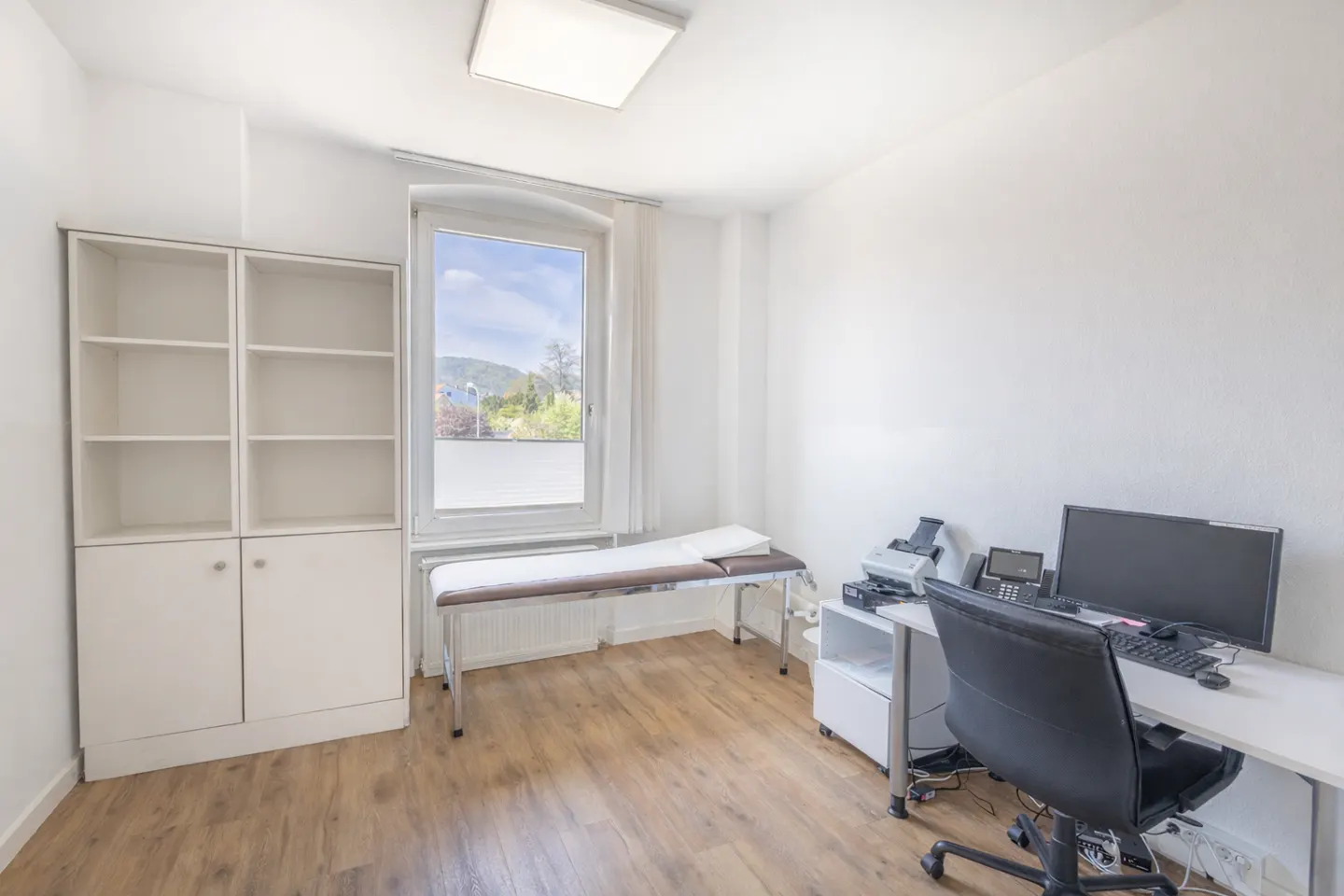 Bright, white doctor's office with a bed, desk, computer, and a view of trees through the window.