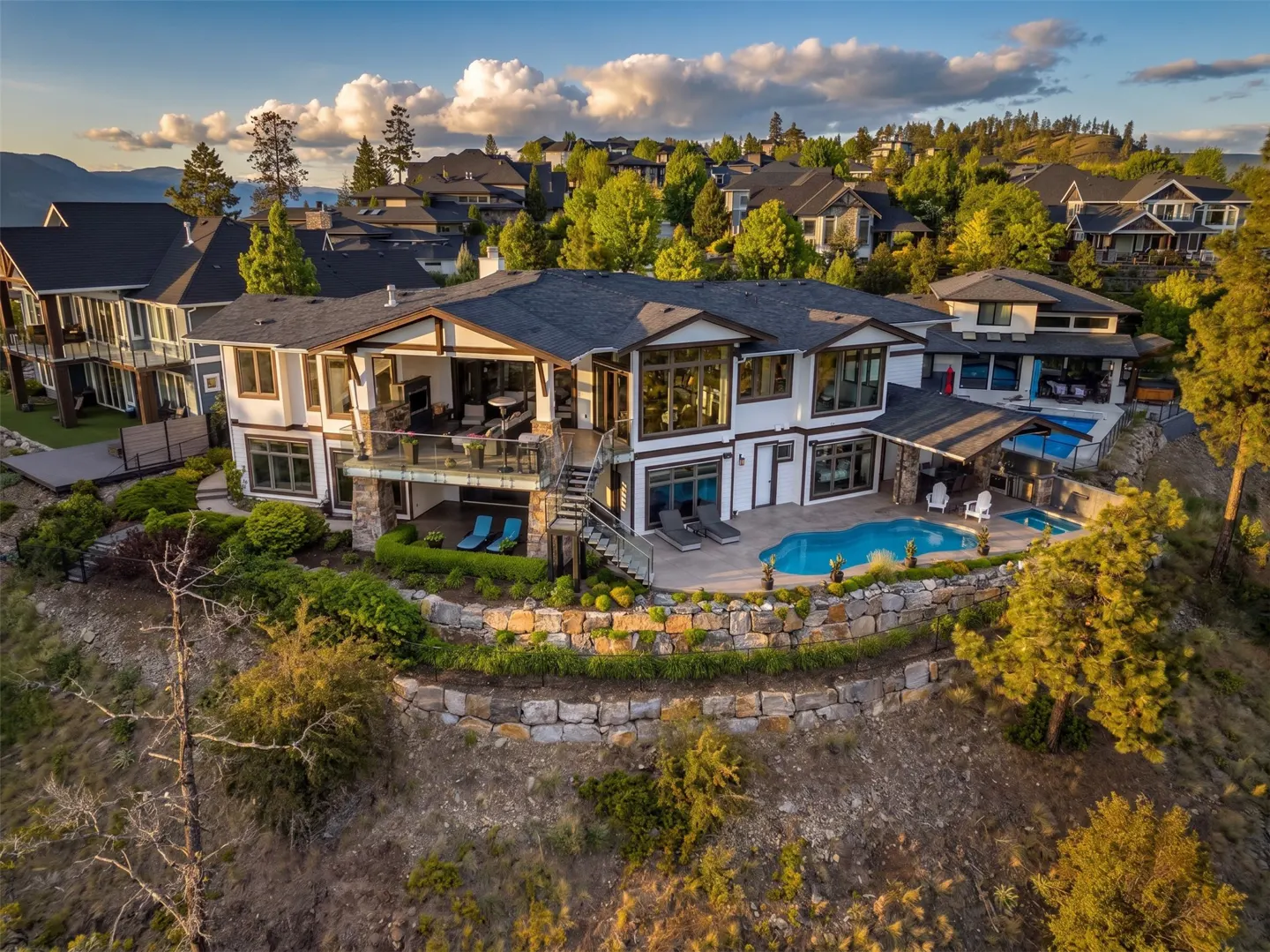 Aerial view of a modern, multi-level white house with a pool, surrounded by lush greenery and other homes on a hillside.