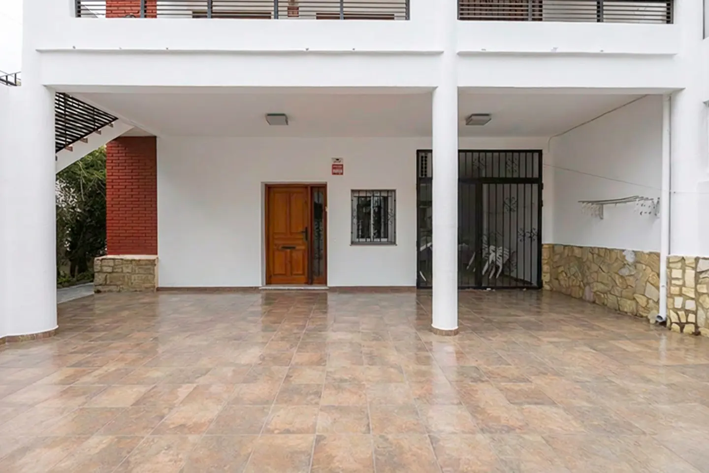 Covered parking area of a white building with a brown tiled floor, a wooden door, and a black metal gate.