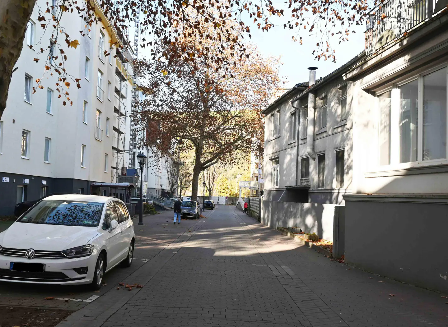 Street view with a white car parked on the left, buildings on both sides, and people walking in the distance.