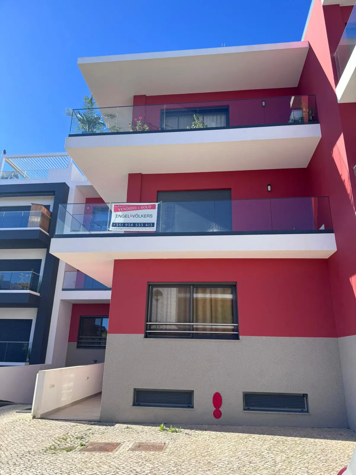 Modern three-story building with red and gray exterior, white balconies with glass railings, and an Engel & Völkers sign.