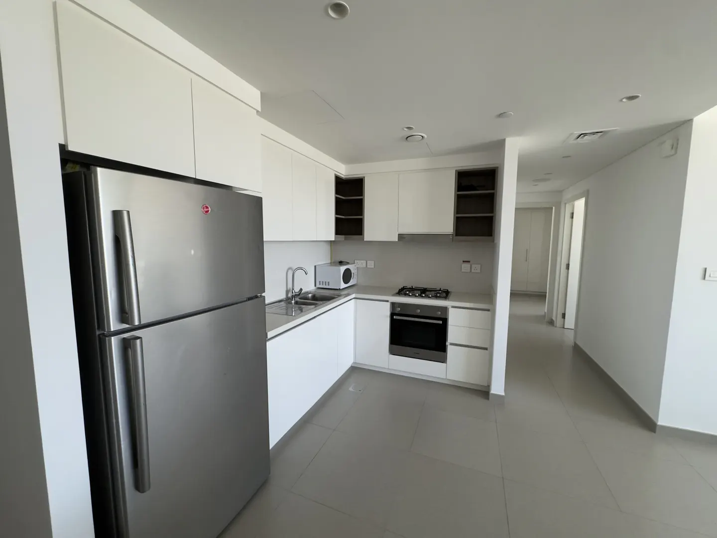 Bright, modern kitchen with stainless steel fridge, white cabinets, microwave, oven, and gas stovetop. Gray tile floor and hallway in background.