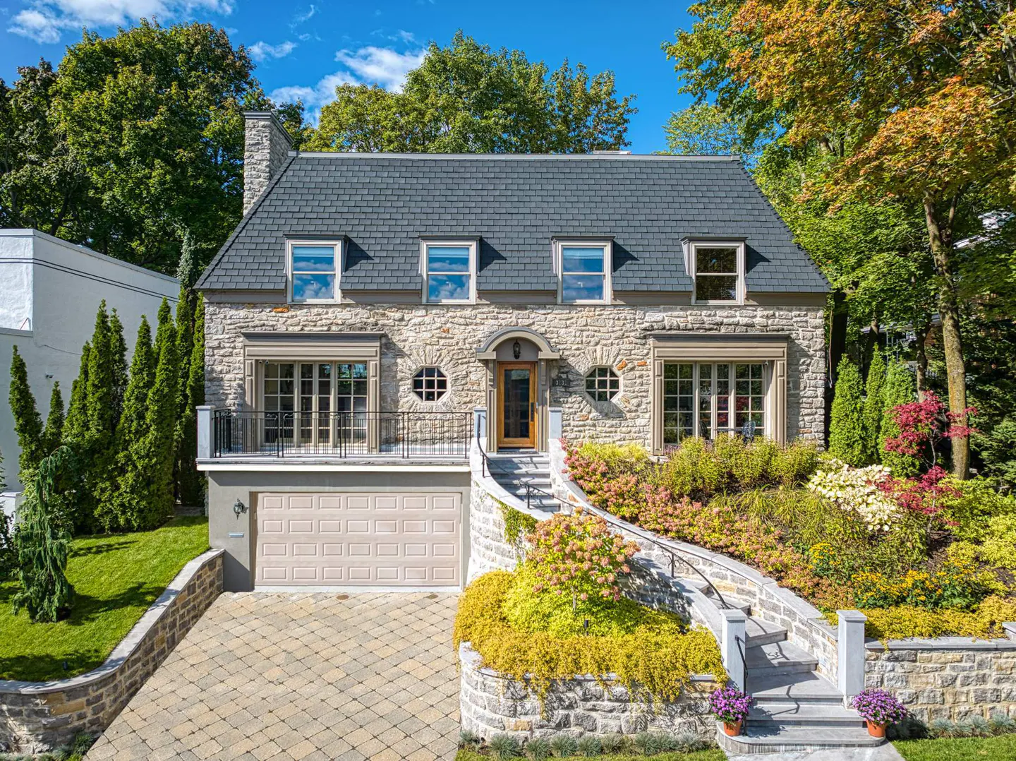 Two-story stone house with a gray roof, dormer windows, and a front yard with flowers and greenery.