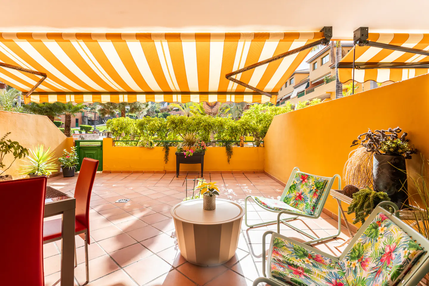A sunny patio with orange walls and a striped awning. Chairs with floral cushions surround a small table. Green plants line the balcony.