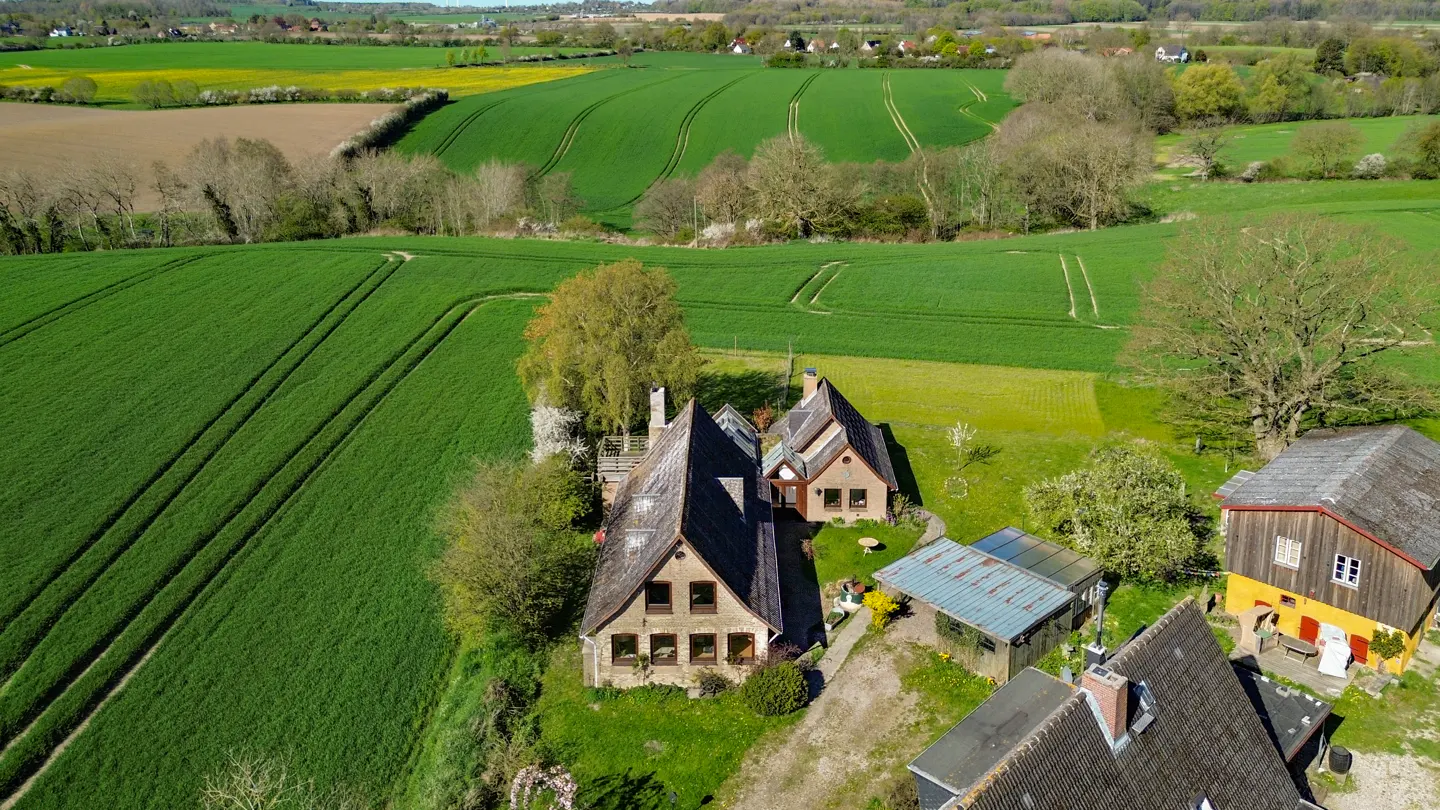 Aerial view of houses with dark roofs surrounded by green fields and trees on a sunny day.