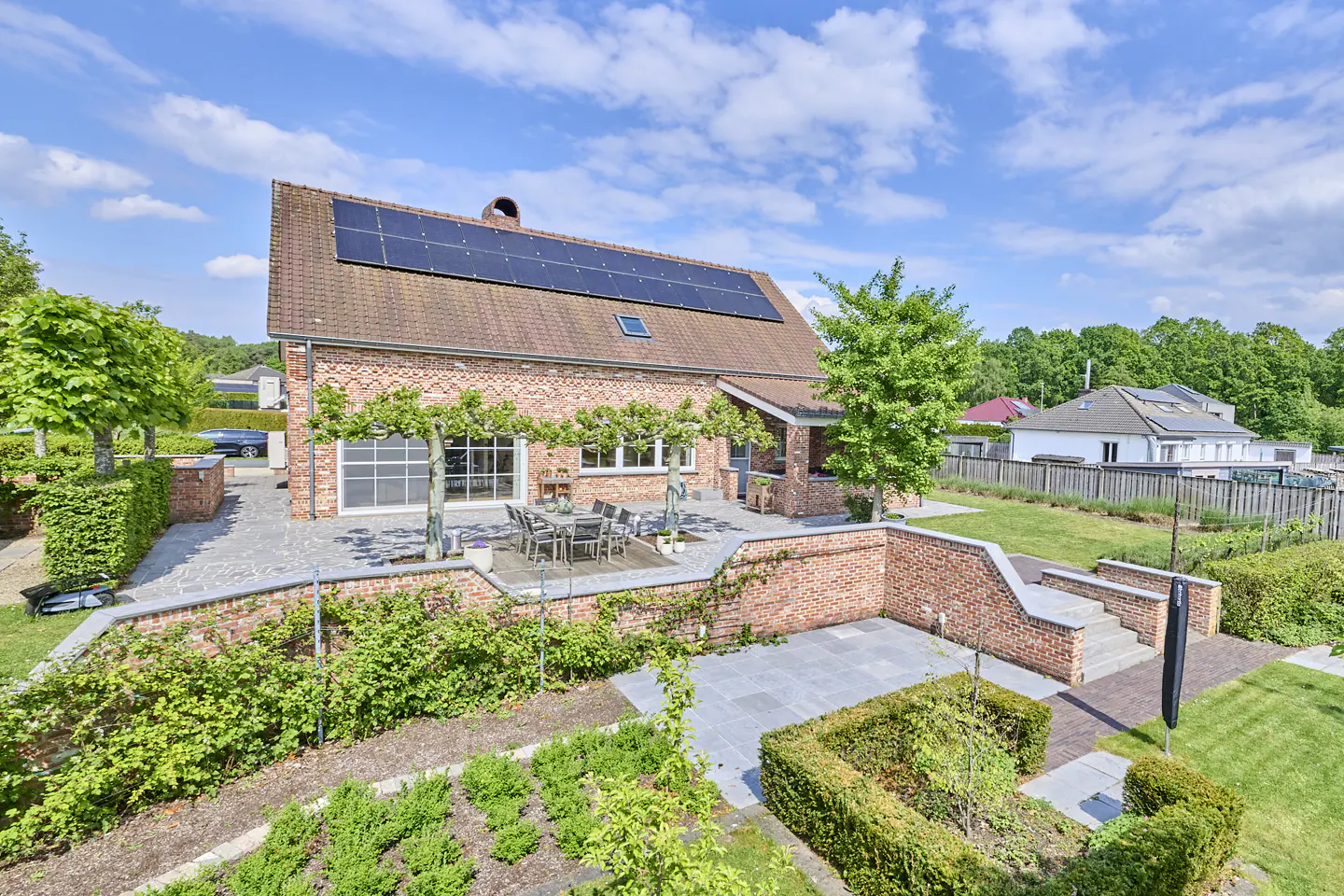 Brick house with solar panels, patio, and garden. Table and chairs on patio. Green trees and blue sky.