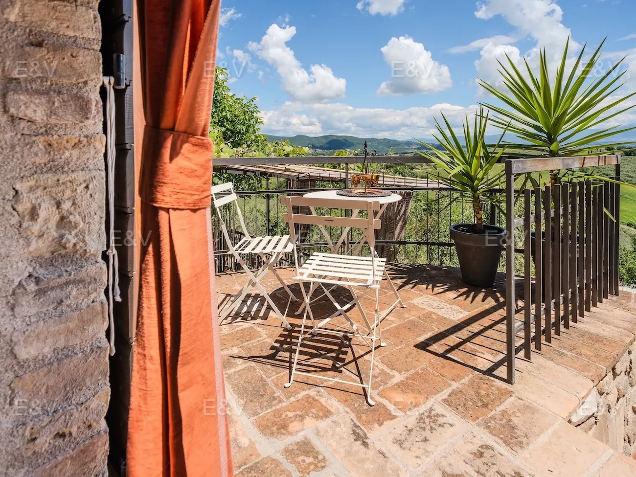 A sunny stone terrace with a white bistro set, potted plants, and a scenic view of rolling hills under a blue sky.
