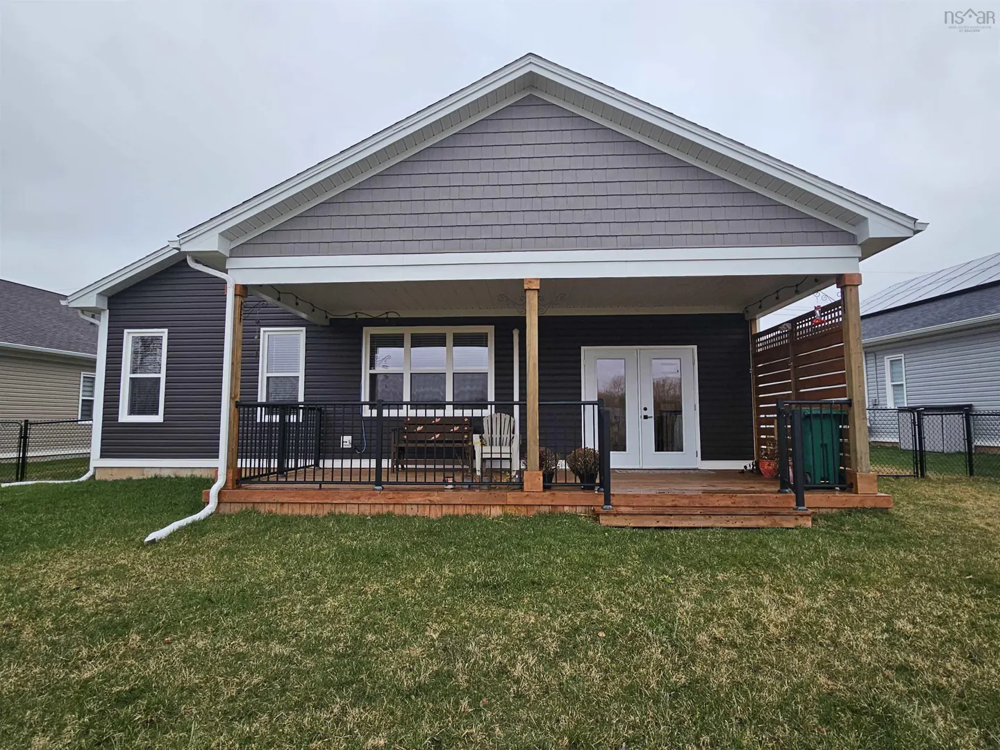 Exterior view of a house with a covered wooden deck, black railings, and a green lawn. The house has gray siding and white trim.