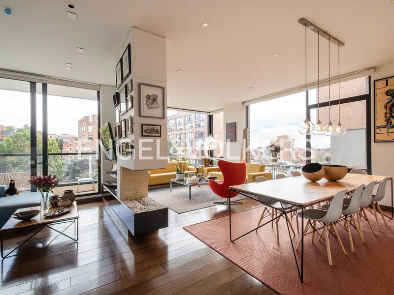 Bright, open-concept living and dining area with wood floors, large windows, and city views. Yellow sofas, red chair, and modern dining table.