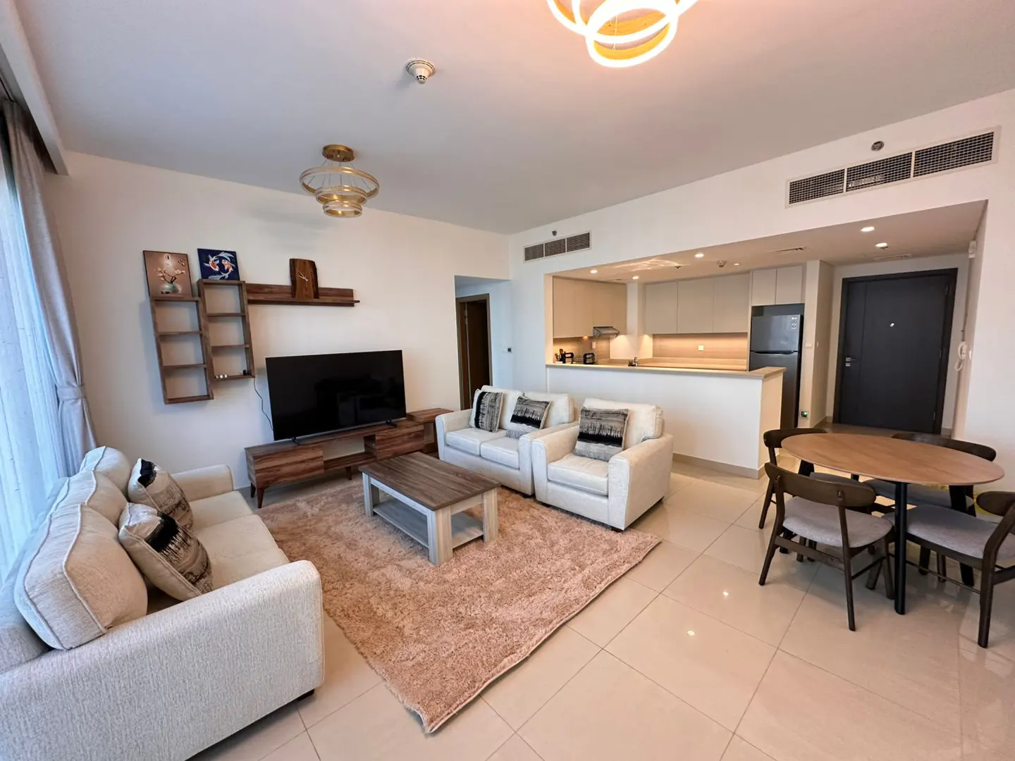 A bright, modern apartment living space with white sofas, a TV, and an open kitchen. A round wooden table with chairs sits near the kitchen area.