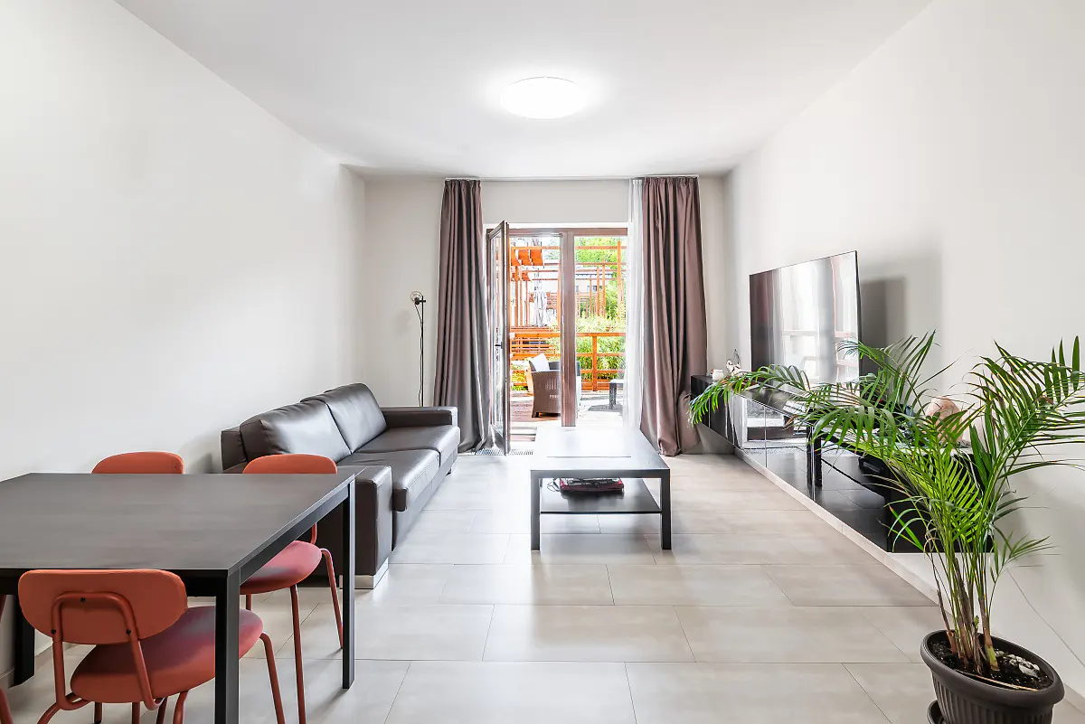 Bright living room with a brown leather sofa, black table and chairs, TV, and a balcony with brown curtains.