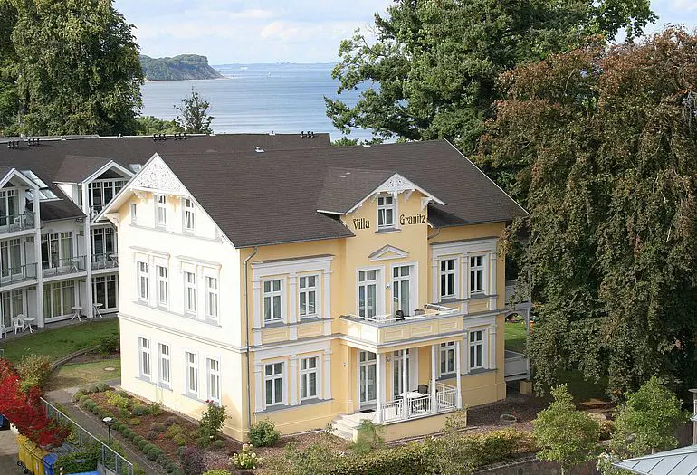 A yellow three-story villa named Villa Granitz with a dark roof, surrounded by trees, overlooking a calm blue sea.