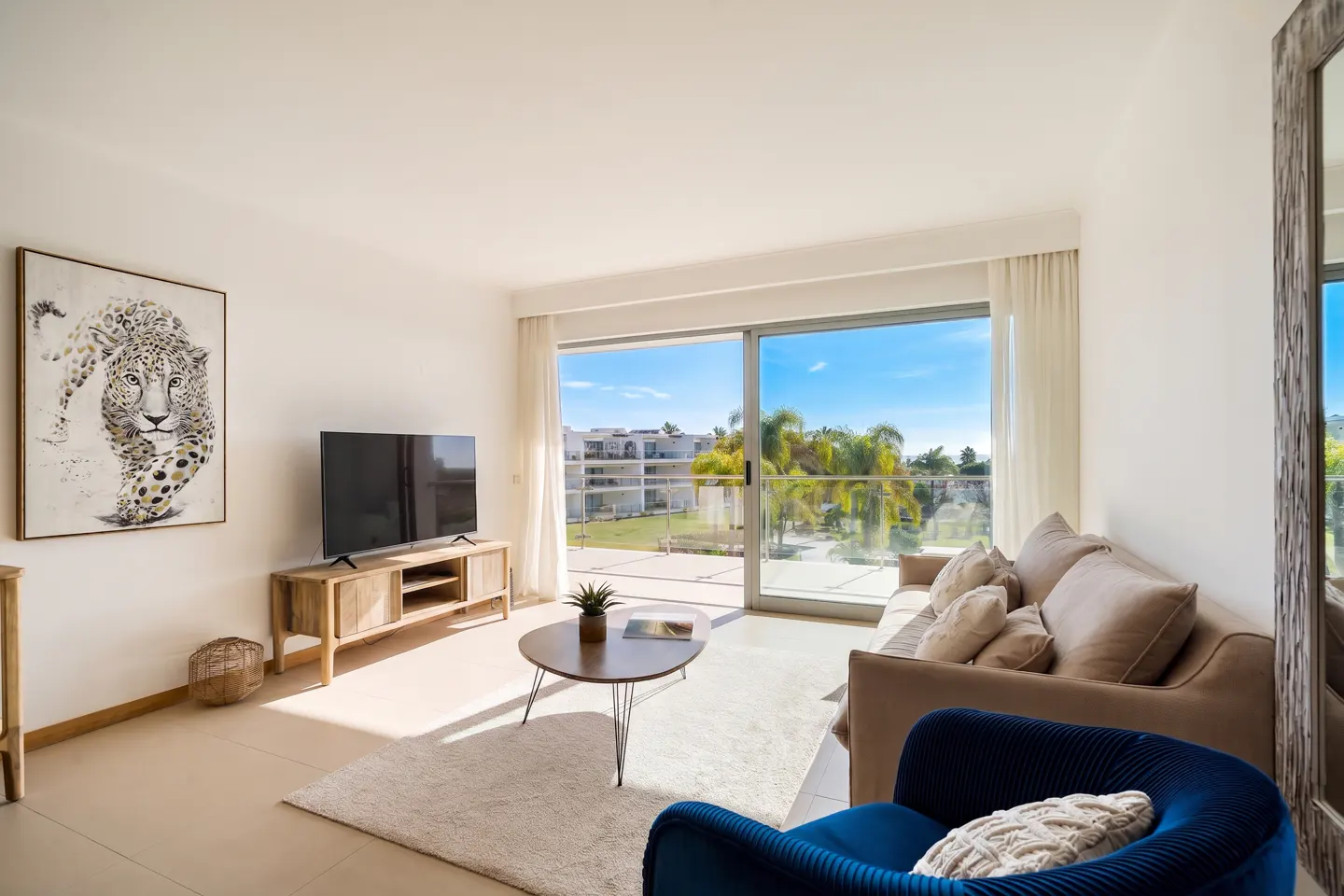 Bright living room with a beige sofa, blue chair, and TV. A large window shows a sunny outdoor view. A leopard print hangs on the wall.