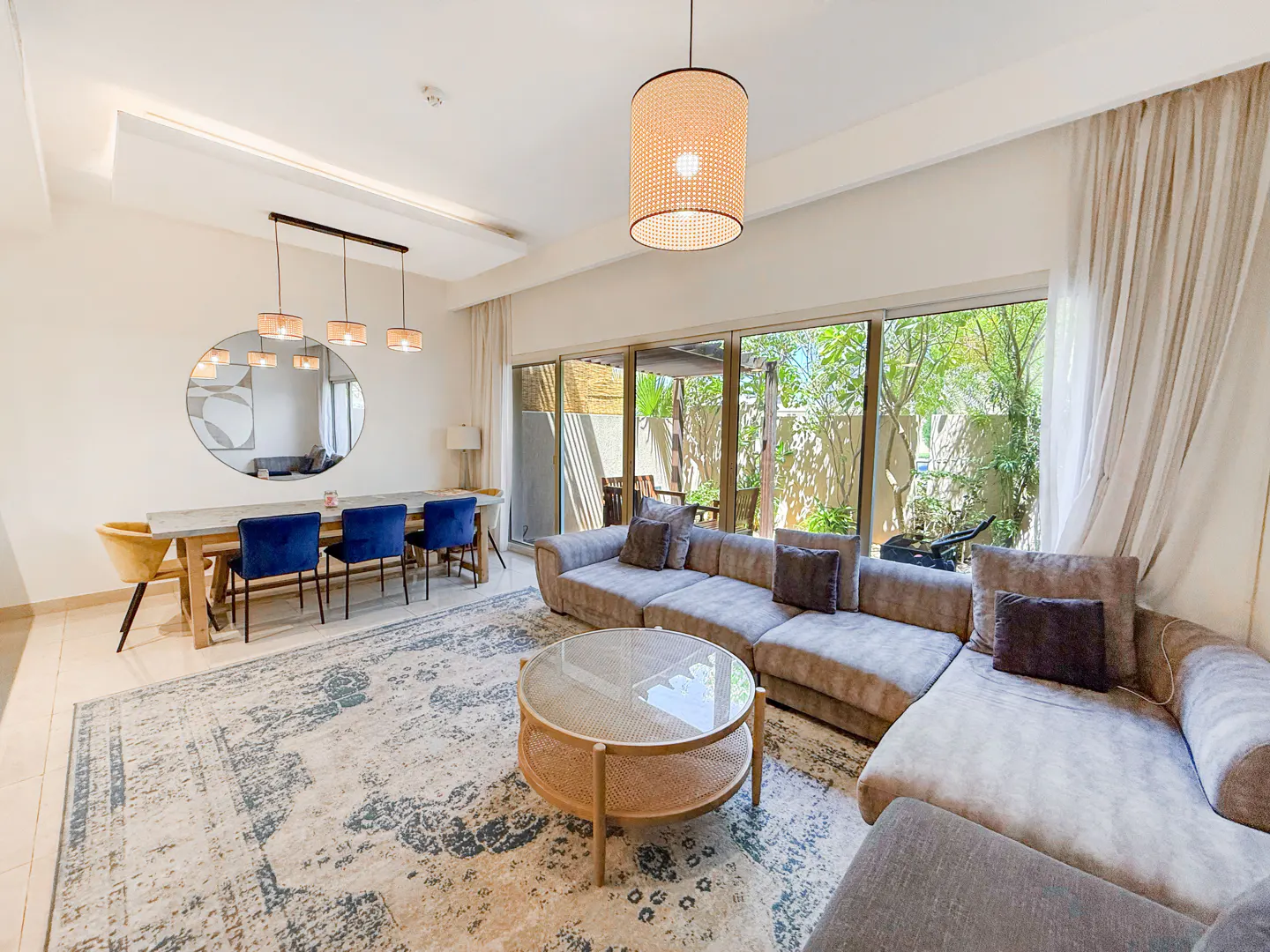 A bright living room with a dining table, blue chairs, a large sofa, and a round coffee table on a patterned rug.