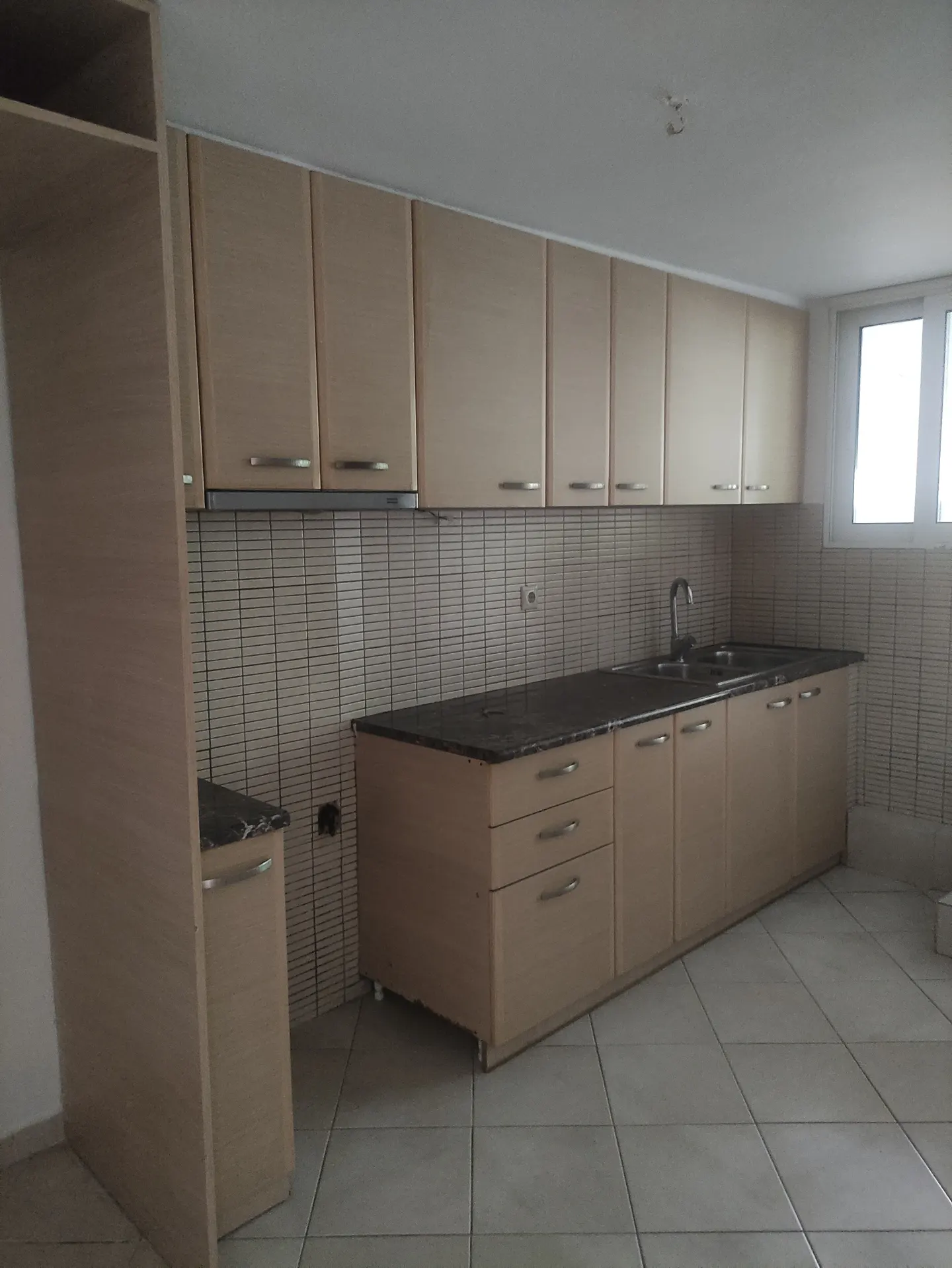 A kitchen with light wood cabinets, dark countertops, and a tiled backsplash. A window is visible in the background.