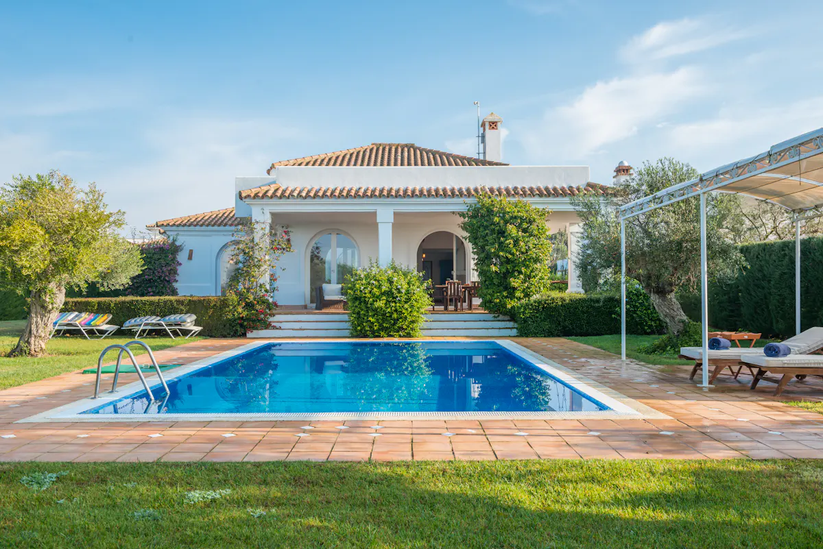 A sunny view of a white villa with a blue swimming pool and lounge chairs.