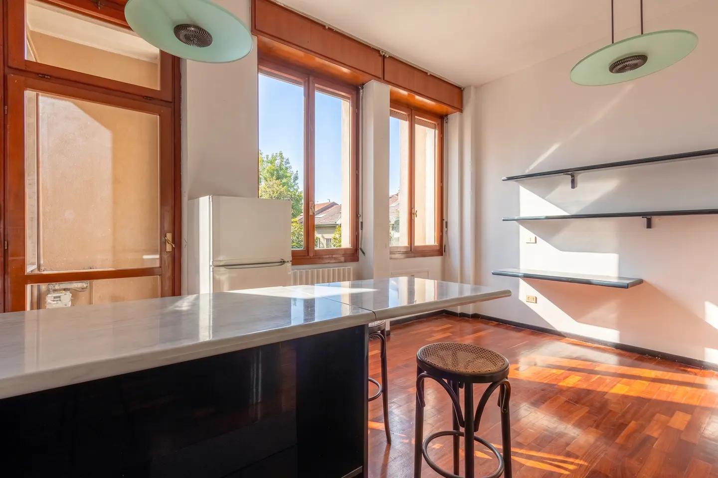 Bright kitchen with wood floors, white walls, and natural light. Features a marble countertop, white fridge, and open shelving.