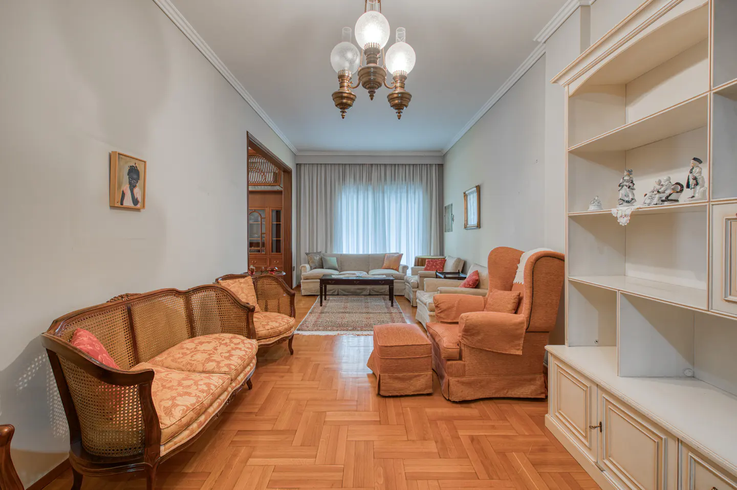 A living room with a sofa, armchair, and a bookshelf. The walls are white, and the floor is wooden. A chandelier hangs from the ceiling.