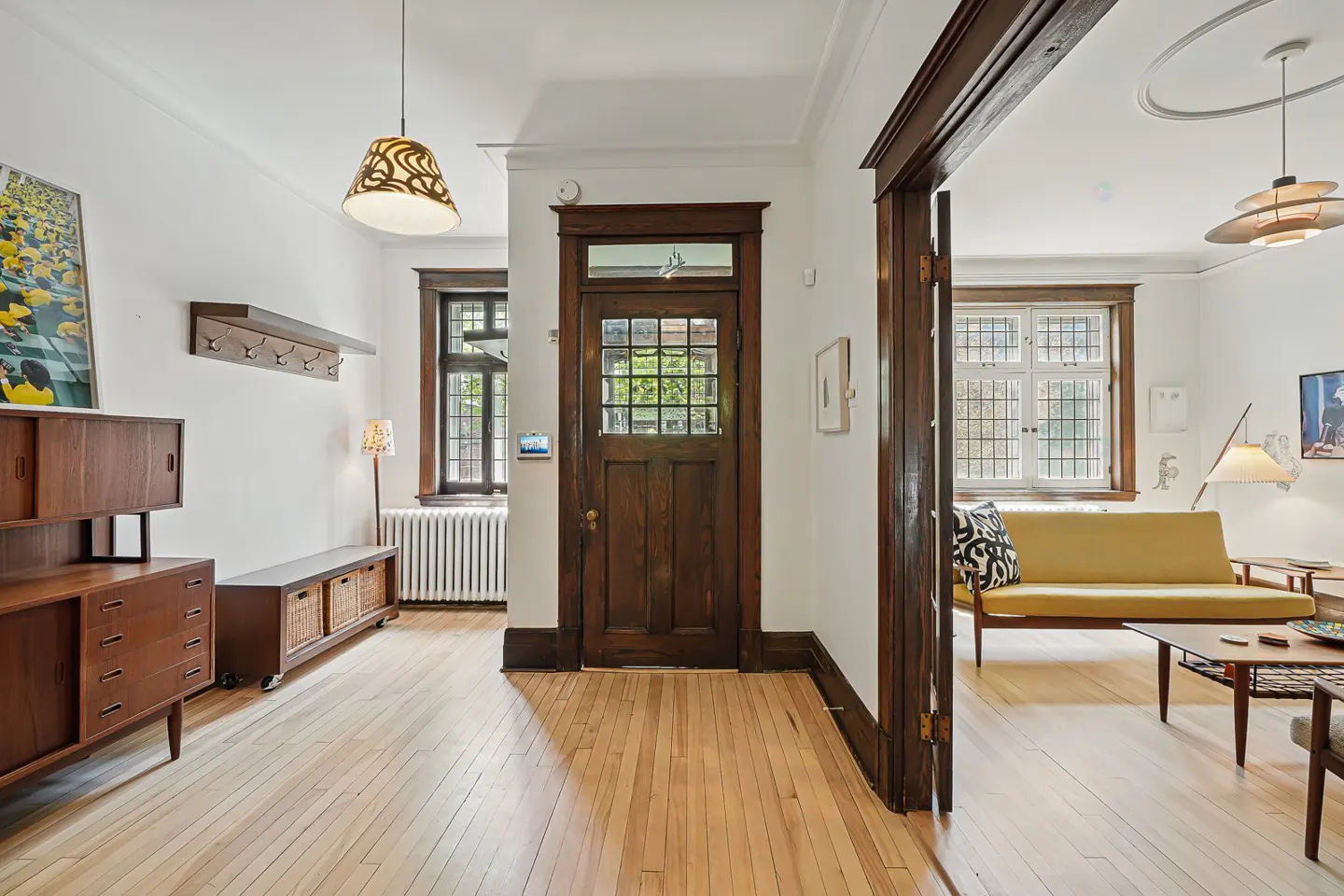 Interior view of a home's entryway with wood floors, a dark wood door, and a glimpse into a living room with a yellow sofa.