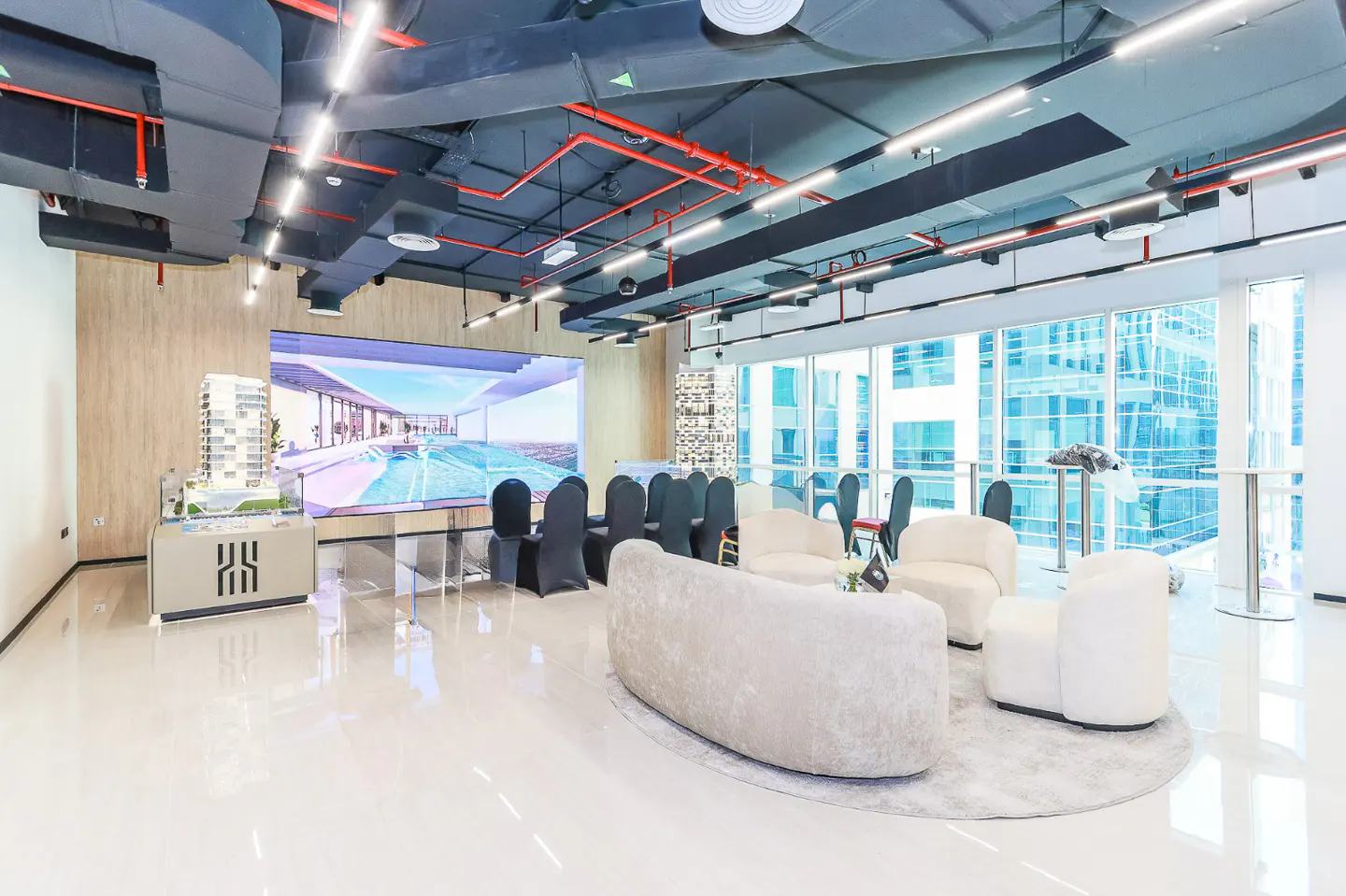Bright, modern real estate showroom with white seating, a building model, and a large screen displaying architectural renderings.