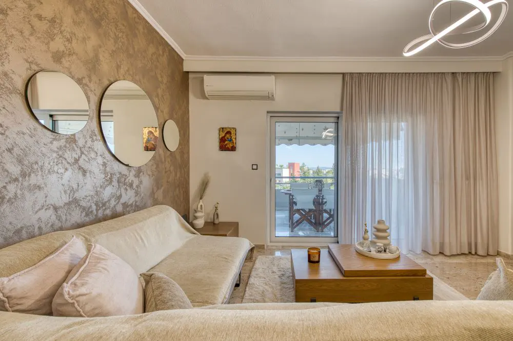 Living room with beige sofa, wood coffee table, and balcony view. Three round mirrors hang on a textured wall. A modern light fixture is on the ceiling.
