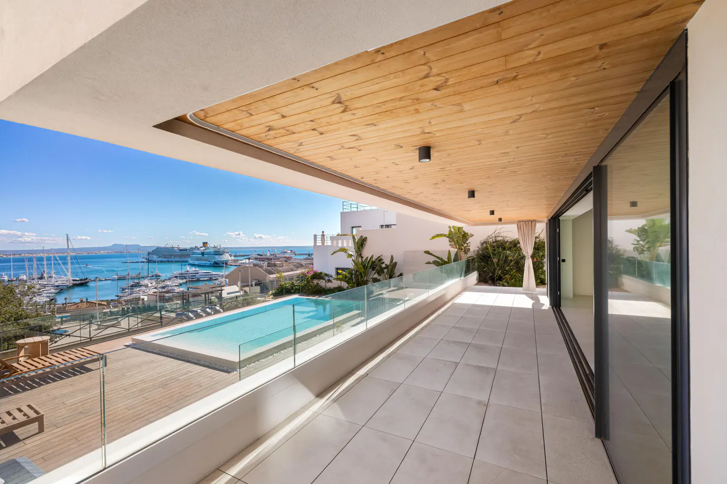Balcony view with a pool, harbor, and cruise ships. The ceiling is wood, and the floor is tiled. Glass railings surround the pool area.