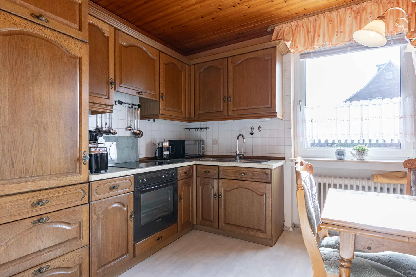 Traditional kitchen with wood cabinets, white tiled backsplash, built-in oven, and a small dining area by the window.