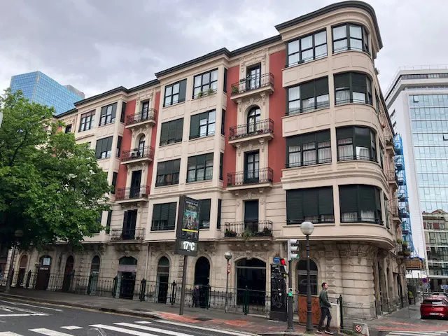 Exterior view of a multi-story building with red and beige facade, black window shutters, and small balconies. Pedestrians walk on a crosswalk.