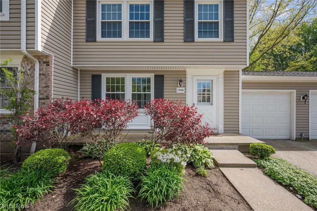 Tan townhouse with black shutters, white door, and garage. Red and green bushes line the front. Concrete walkway leads to the entrance.
