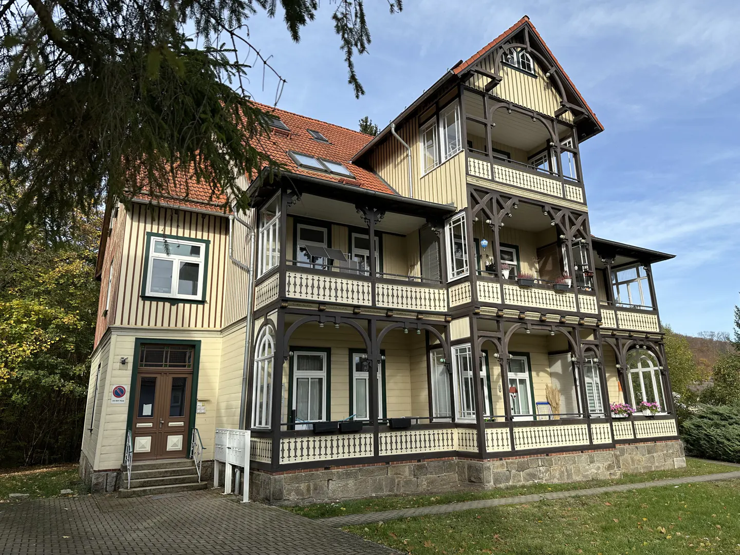 A three-story yellow house with brown trim and a red roof, featuring balconies and a stone foundation.