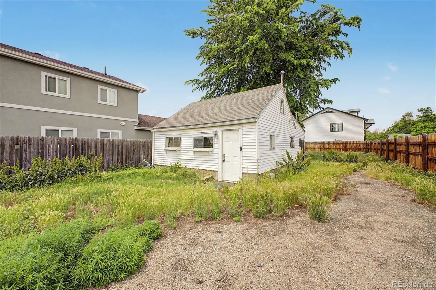 Small white detached garage in a backyard with overgrown grass, a gravel path, and wooden fencing under a blue sky.