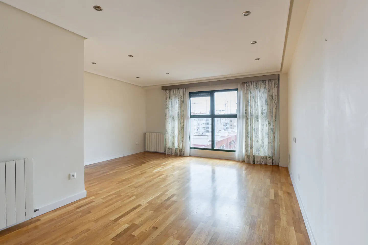 Bright, empty room with hardwood floors, white walls, and a large window with floral curtains overlooking a city.