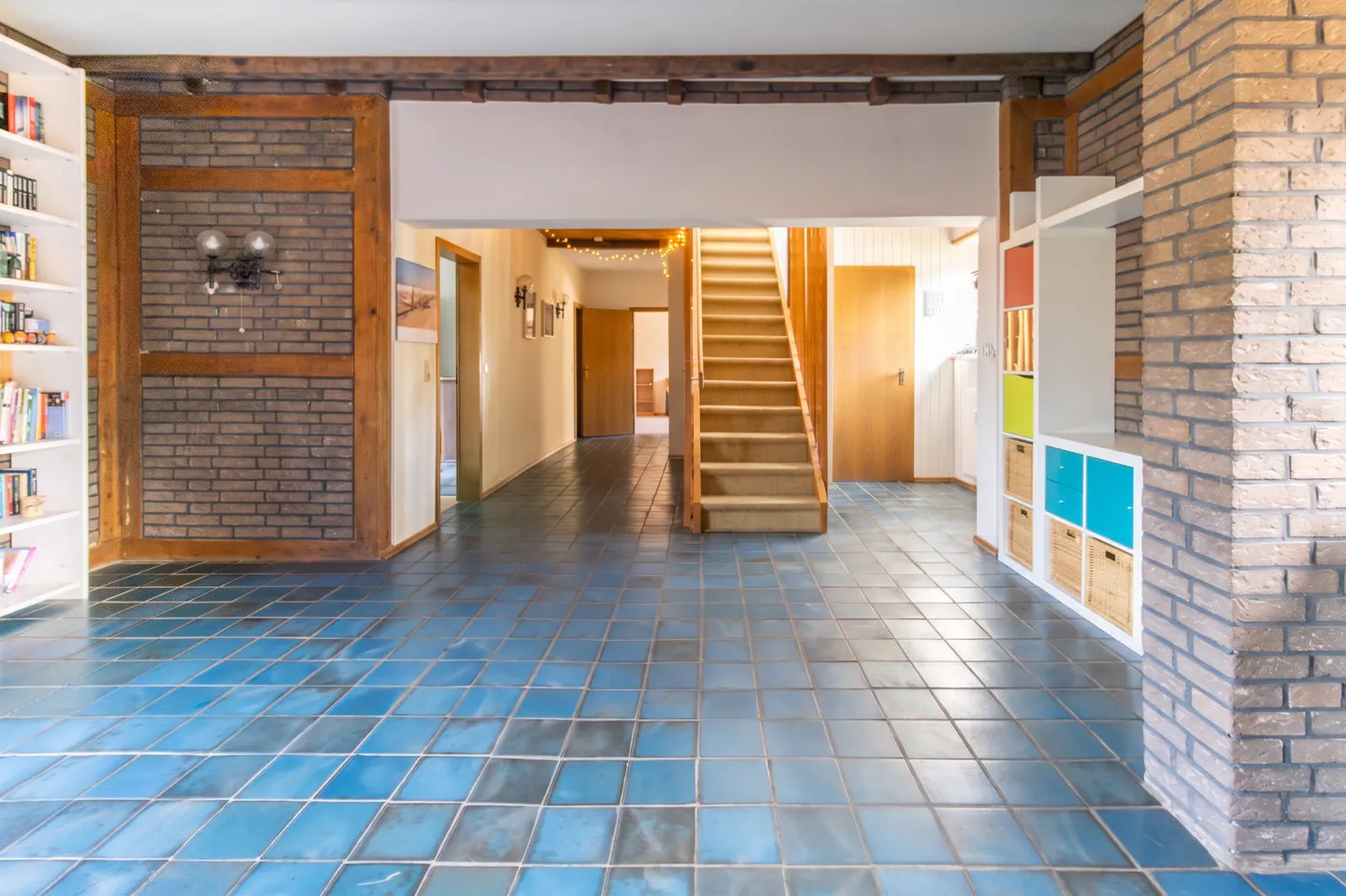Interior view of a home's entryway with blue tile flooring, brick walls, wooden beams, staircase, and built-in shelving.
