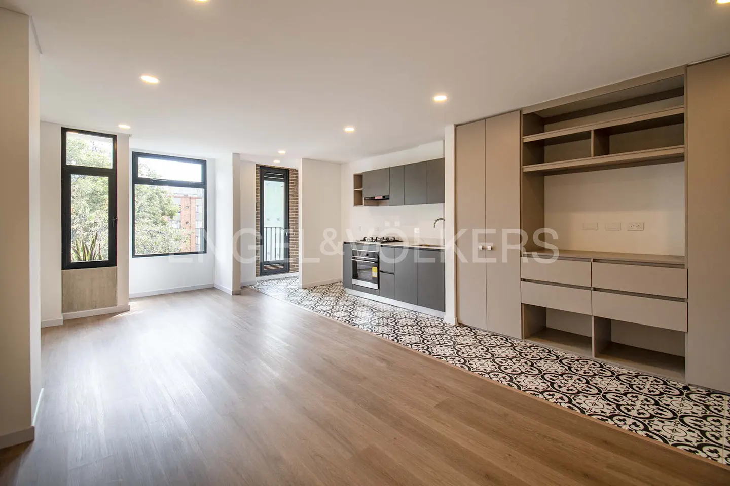Bright, modern apartment interior with wood floors, gray kitchen cabinets, and a black and white patterned tile floor. Large windows offer natural light.