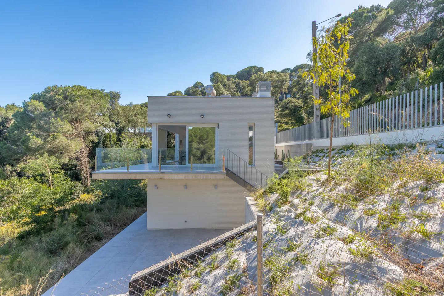 Modern beige house with a glass balcony, set on a sloped, wooded hillside under a clear blue sky.