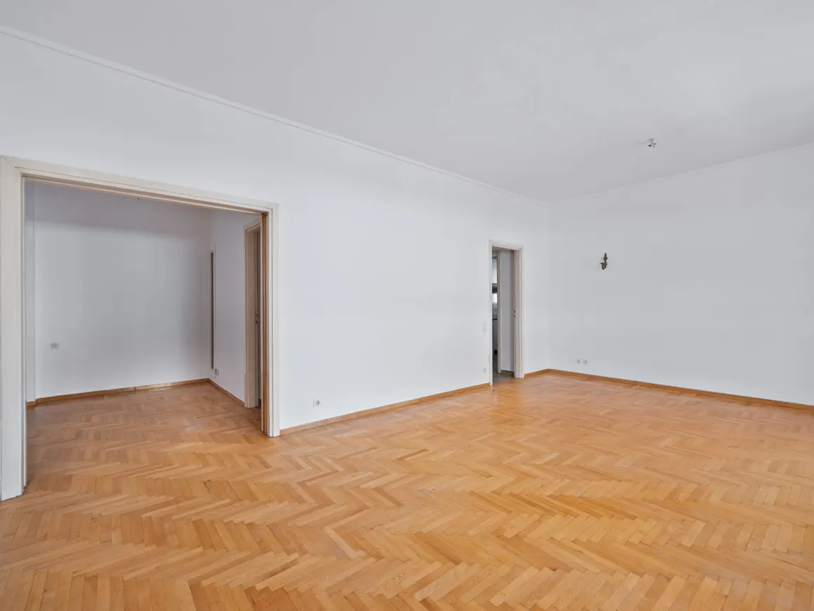 Empty room with white walls, herringbone wood floor, and two doorways.
