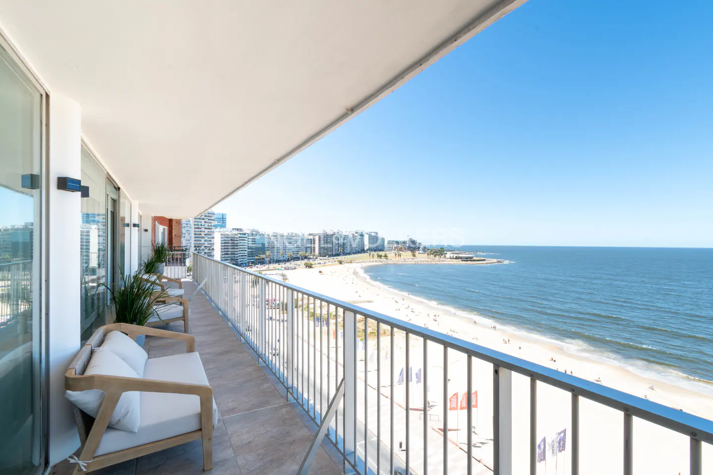 Balcony view of a sandy beach and ocean. White sofas and chairs line the balcony. City buildings are visible in the background.