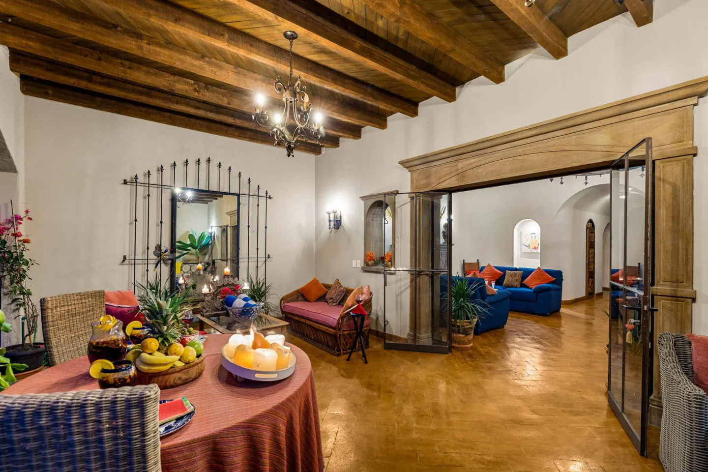 Interior view of a living room with a table, fruit, candles, and blue sofas in the background. The room has wooden beams and a chandelier.
