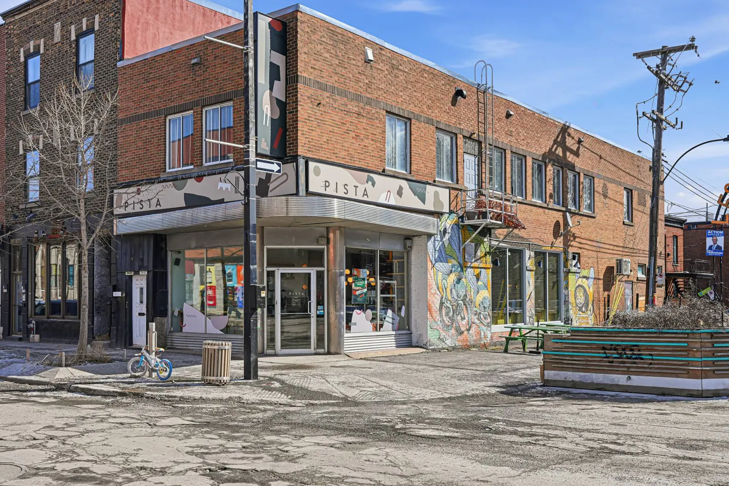 Exterior view of Pista, a brick building with large windows and a mural. A blue bicycle sits on the sidewalk.