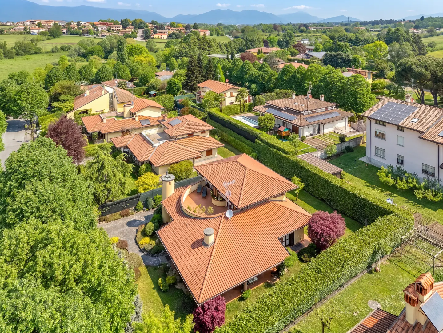 Aerial view of a luxury home with a terracotta roof, surrounded by green trees and manicured hedges in a suburban neighborhood.