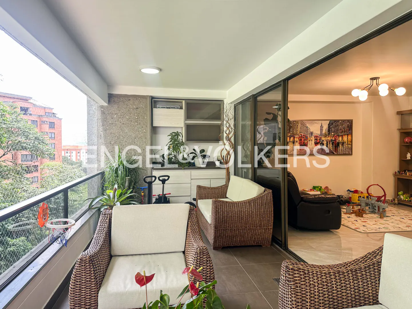 A bright balcony with wicker furniture and a view of trees and buildings. Sliding glass doors lead to a living room with toys and a London scene painting.