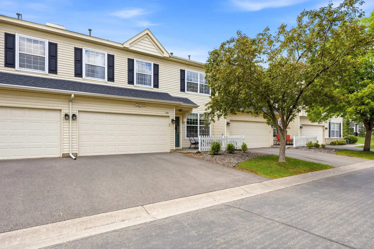 Beige townhouses with black shutters and white garage doors line a street under a blue sky with green trees.