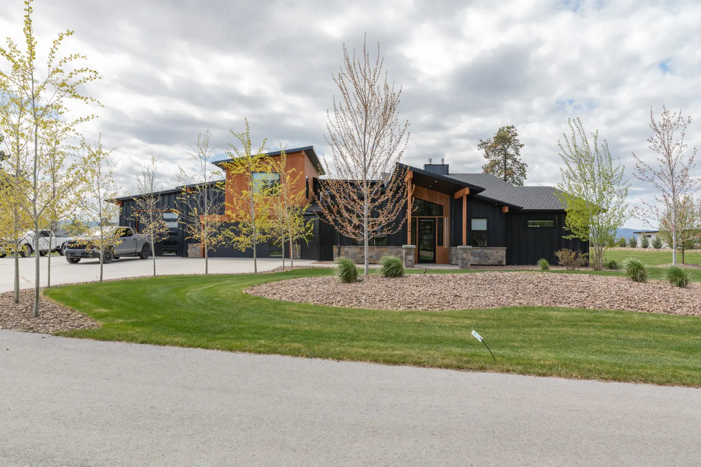 Modern black house with wood accents, a stone base, and a circular driveway with green grass and small trees.