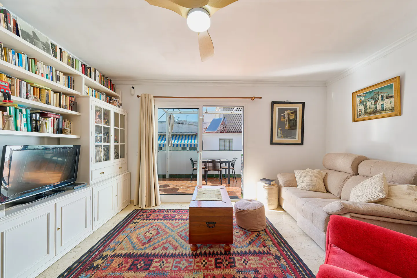 Bright living room with white walls, a beige sofa, and a red armchair. A large bookcase is filled with books. A sliding door leads to a balcony.
