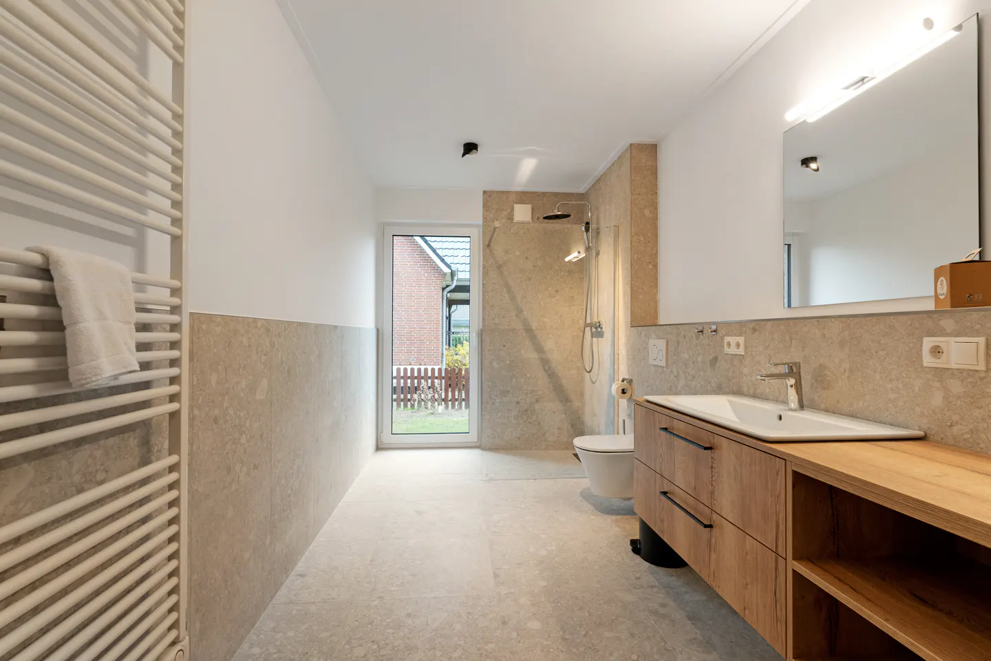 Bright bathroom with beige stone walls, a wood vanity, and a white towel warmer. A shower and toilet are visible.