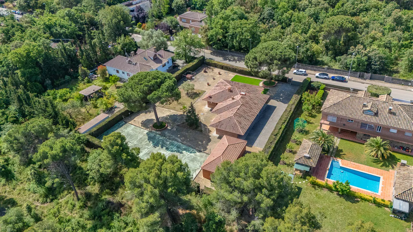 Aerial view of a large property with a red-tiled roof house, tennis court, pool, and lush green trees.